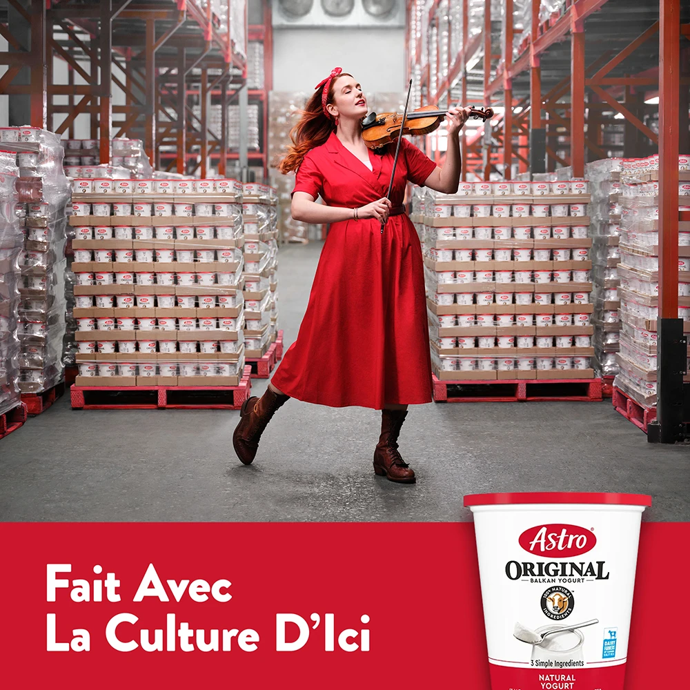 Une violoneuse en robe rouge joue du violon entre des palettes de yogourt Astro Original.